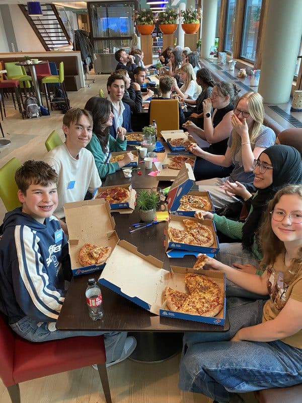 Leerlingen aan een lange tafel met pizzadozen, lachend naar de camera